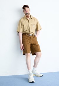 Helles beige Kurzarmhemd mit zwei Brusttaschen, braune Shorts und weiße Sneakers mit grauen Akzenten. Das Model steht vor einer weißen Wand.