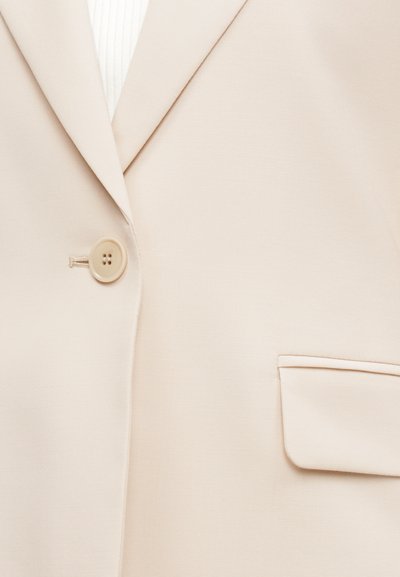 Μπεζ σακάκι με ένα κουμπί, γιακά με εγκοπή και τσέπη με καπάκι στην εμπρόσθια δεξιά πλευρά.