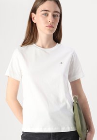 T-shirt bianco in cotone con scollatura rotonda e maniche corte. Presenta un piccolo design ricamato sul petto. Jeans neri e una borsa verde.
