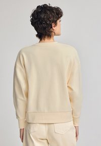 Felpa beige chiaro con vestibilità rilassata, polsini e orlo a coste. Tessuto morbido con un design semplice e pulito. La vista posteriore mostra uno stile minimalista.