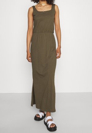 Robe maxi vert olive avec un décolleté carré, une taille froncée et une fente latérale, associée à des sandales blanches avec une semelle épaisse noire.