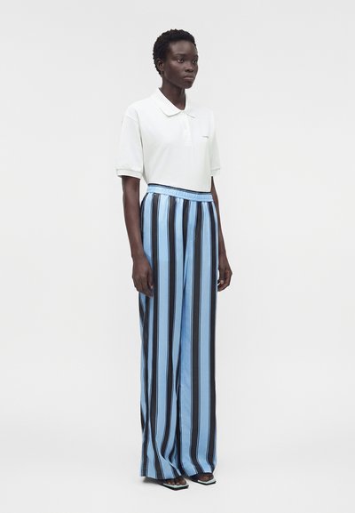 Άτομο όρθιο που φοράει λευκό πόλο με κοντά μανίκια συνδυασμένο με ψηλόμεση παντελόνα σε κάθετες μπλε και μαύρες ρίγες.