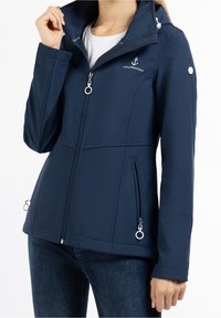 Marinblå mjukshelljacka med hög krage, dragkedjefickor och metalliska detaljer; slank design med en jämn textur.