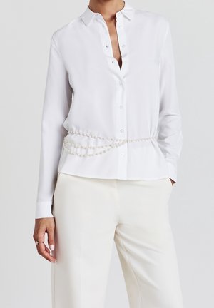 Vrouw draagt een witte blouse met lange mouwen en knopen, een drielaagse parelriem en crèmekleurige broek, één hand in de zak en één ontspannen.