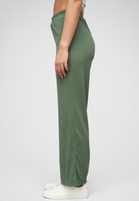 Grønne bukser med elastisk midje, ribbet tekstur og sideslitser, sammen med hvite joggesko. Designet er løst og rett i bena.