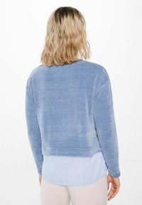 Lichtblauwe gebreide sweater met een relaxte pasvorm, ribbelstructuur en verlaagde schouders, gelaagd over een lichtblauw gestreept shirt.