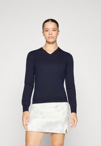 Marinblå stickad tröja med v-ringning, ribbade muddar och nederkant. Bärs med en ljus mönstrad kjol, vilket framhäver tröjans figurnära design.