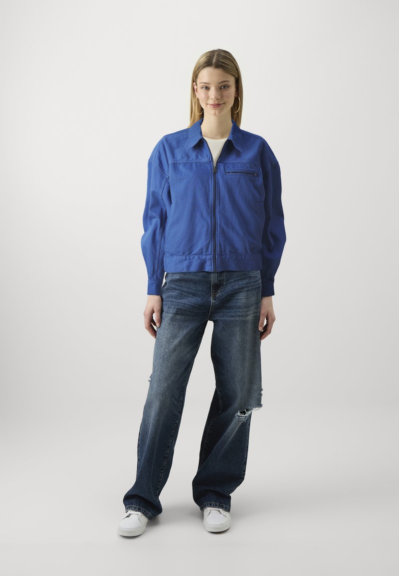 Blaue Baumwoll-Bomberjacke mit einem Frontreißverschluss, seitlichen Taschen und einer Brusttasche, kombiniert mit abgetragenen Dunkeljeans und weißen Sneakers.