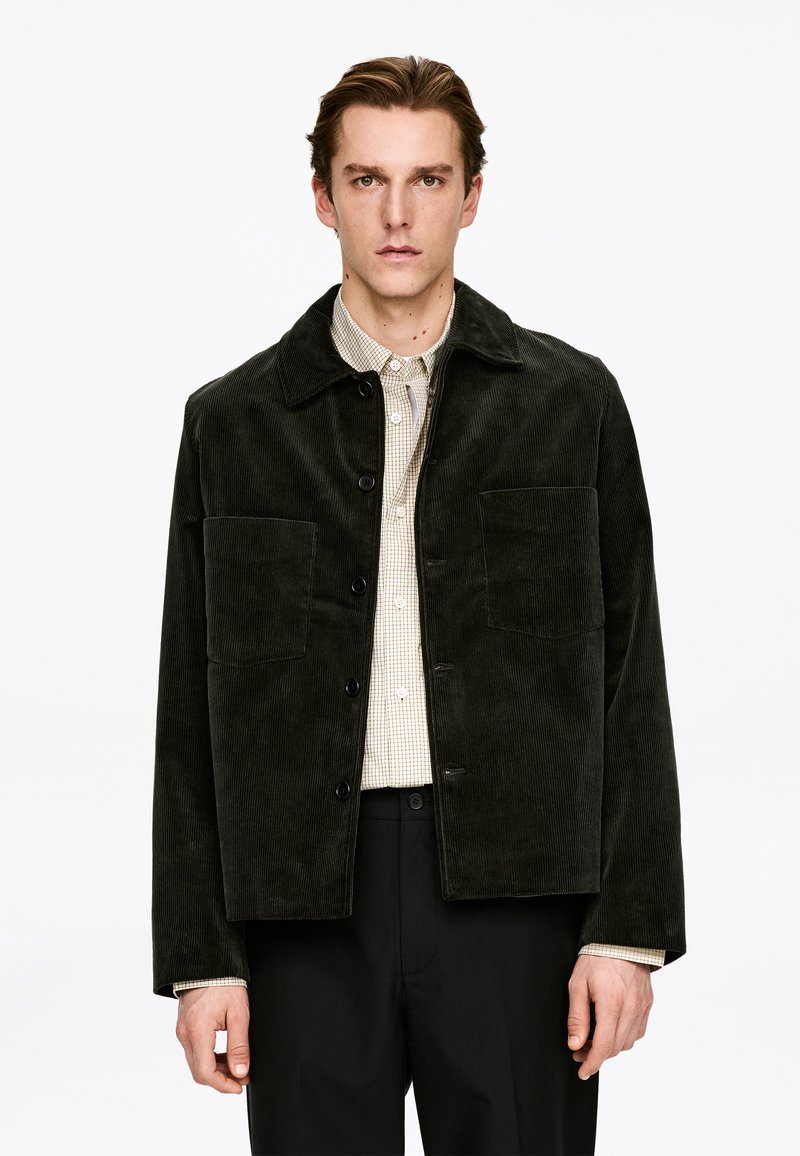 Veste en velours côtelé vert foncé avec fermeture à boutons, deux poches avant et un col, portée sur une chemise à carreaux claire.