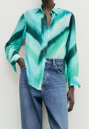 Person iført løs teal og grøn tie-dye skjorte med knapper, stukket ind i højtaljede blå jeans, stående med den ene hånd i lommen.