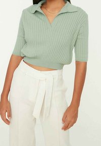 Crop top polo en maille côtelée vert clair avec manches courtes, associé à un pantalon blanc taille haute avec ceinture à nouer.