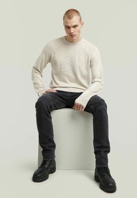 Helle beige Strickpullover mit strukturierten Mustern, kombiniert mit schwarzen, distressed Jeans und schwarzen Schnürstiefeln, präsentiert vor einem neutralen Hintergrund.