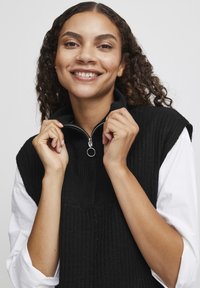 Schwarzer, gerippter ärmelloser Pullover mit hohem Kragen und halbem Reißverschluss. Modell lächelt und hält den Reißverschluss; weißes Hemd darunter sichtbar.