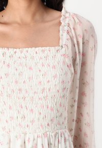 Rochie florală cu un corset plisat, mâneci din dantelă semi-transparente, material de culoare crem deschis și flori mici roz distribuite uniform. Detaliu cu volan la guler.