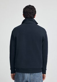 Marinblå sweatshirt med hög krage, tillverkad av ett strukturerat tyg. Har långa ärmar och en avslappnad passform som sträcker sig till midjan.