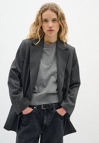 Mørkegrå blazer med en struktureret design, lagd over en lys grå sweater. Sorte højtaljede jeans med en afslappet pasform og et bælte.
