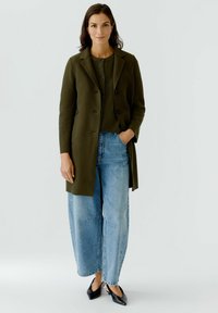 Manteau en laine vert olive au design ajusté, avec des boutons noirs, associé à un pull texturé vert foncé et un jean taille haute bleu clair. Chaussures noires.