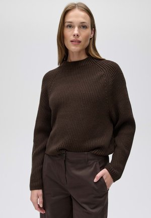 Vrouw met een lichte huid en steil lichtbruin haar draagt een donkerbruine gebreide trui en donkerbruine broek, staand voor een effen lichte achtergrond.