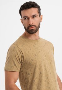 Tan kortærmet t-shirt lavet af blødt materiale, med et subtilt mønster af bogstaver og symboler gennem hele stoffet. Rund halsudskæring.