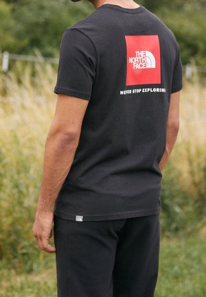Homme portant un t-shirt noir North Face avec un carré rouge et un logo blanc avec le texte « Never Stop Exploring », debout à l'extérieur près de l'herbe.