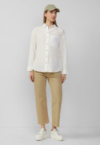 Witte linnen overhemd met lange mouwen en een borstzak, gecombineerd met beige cropped broek en witte sneakers; lichtgroene pet.