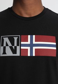 Schwarzes T-Shirt mit kurzen Ärmeln, das ein graues Feld mit einem schwarzen Buchstaben "N" und eine horizontale norwegische Flagge in rot, weiß und blau zeigt.