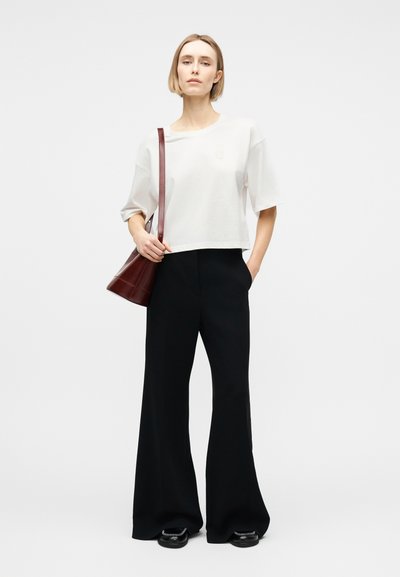 Γυναίκα στέκεται φορόντας λευκό, άνετο τοπ με κοντά μανίκια, μαύρο παντελόνι φαρδύ στο κάτω μέρος, μαύρα παπούτσια και σκούρα καφέ τσάντα ώμου.