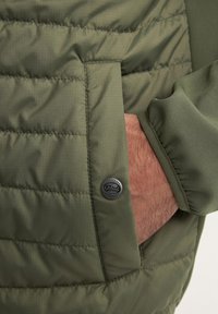 Giacca imbottita verde oliva con texture trapuntata, dotata di una tasca laterale e dettaglio del bottone a pressione nero con marchio. Tessuto morbido e polsini aderenti.