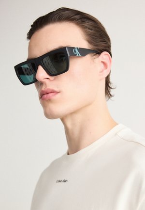 Black square sunglasses with dark lenses and light blue "CK" logo on the arm, worn by a model with short hair, featuring light-colored shirt.