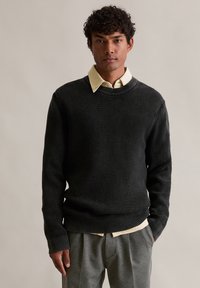 Černý žebrovaný pletený svetr s kulatým výstřihem, přes světlou žlutou košili s límečkem. Nošený s šedými kalhotami, což ukazuje na volný styl.