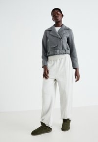 Chaqueta de moto de lana gris con detalles de cremallera, cintura ceñida, pantalones jogger gris claro y botas de tobillo verde oliva con una textura suave.