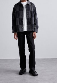 Schwarz-grau kariertes Hemdjacke mit Kragen, weißes T-Shirt darunter, kombiniert mit schwarzen Hosen und schwarzen Schuhen. Glatte Stofftextur.