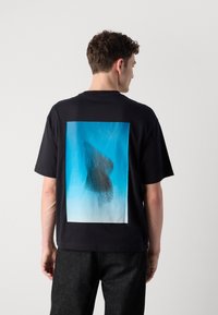 Fekete, rövid ujjú póló, nagy kék és fehér grafikai nyomattal a hátsó részén, amely textúrált sötét folt mintázattal rendelkezik.