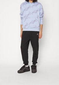 Hellblaue Kapuzenjacke mit lila grafischem Text, schwarze Jogginghosen mit Jordan-Logo und schwarze Sneaker. Das Modell steht in neutraler Pose.