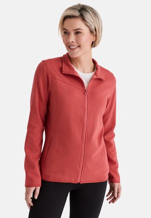 Woman smiling, wearing a red zip-up fleece jacket over a white shirt and black pants, standing against a plain light background.