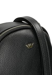 Borsa in pelle nera con texture, dotata di una cerniera argentata e di un piccolo emblema dorato con un design simile a delle ali sulla parte frontale.