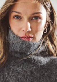 Grauer, flauschiger Rollkragenpullover mit strukturierter Oberfläche. Nahaufnahme eines Gesichts mit leichter Make-up, natürlichem Haar und silbernen Creolen.