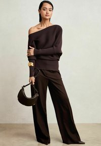 Dunkelbrauner gerippter Strickpullover, der schulterfrei getragen wird, kombiniert mit weit geschnittenen Hosen. Das Modell hält eine schwarze Ledertasche mit goldenen Akzenten.