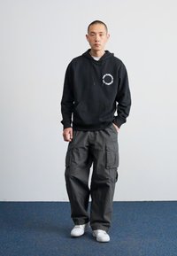 Hoodie noir avec une poche frontale et un logo blanc, associé à un pantalon cargo gris foncé doté de plusieurs poches. Des baskets blanches complètent le look.