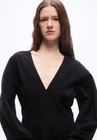 Vrouw met lang bruin haar, gekleed in een zwarte V-hals jurk met lange mouwen, tegen een effen witte achtergrond.