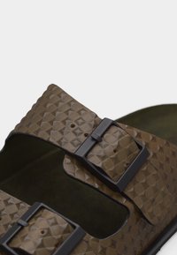 Cinturino del sandalo marrone con motivo a diamante texturizzato e due fibbie nere regolabili su sfondo semplice.