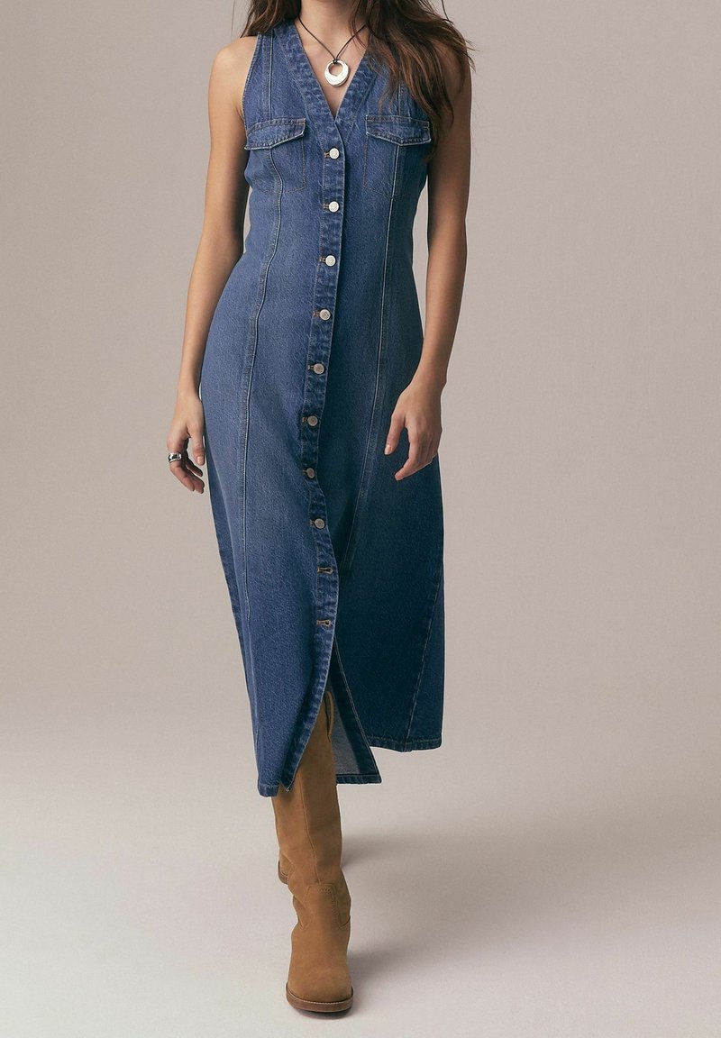 Femme portant une robe en jean bleu sans manches à boutons et des bottes en daim beige, accessoirisée d'un collier et de bagues en argent, marchant vers l'avant.