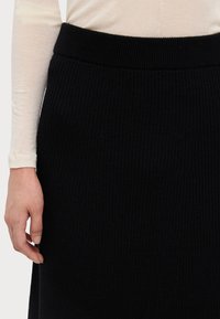 Sort ribbet striknederdel med et bredt, tekstureret taljebånd, vist sammen med en lysefarvet langærmet top, der fremhæver materiale og design.