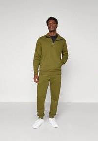 Olivno zelena majica z jopico z zadrgo, kombinirana z usklajenimi trenirkami. Tkanina je mehka, z rebrastimi manšetami in subtilnim logotipom.