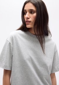 Tricou din bumbac gri, cu guler rotund, mâneci scurte și o croială lejeră, având o textură netedă și detalii de cusătură subtile.