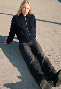 Tânără cu păr blond, purtând o jachetă neagră cu fermoar și blugi închiși la culoare, așezată pe beton la lumina soarelui.