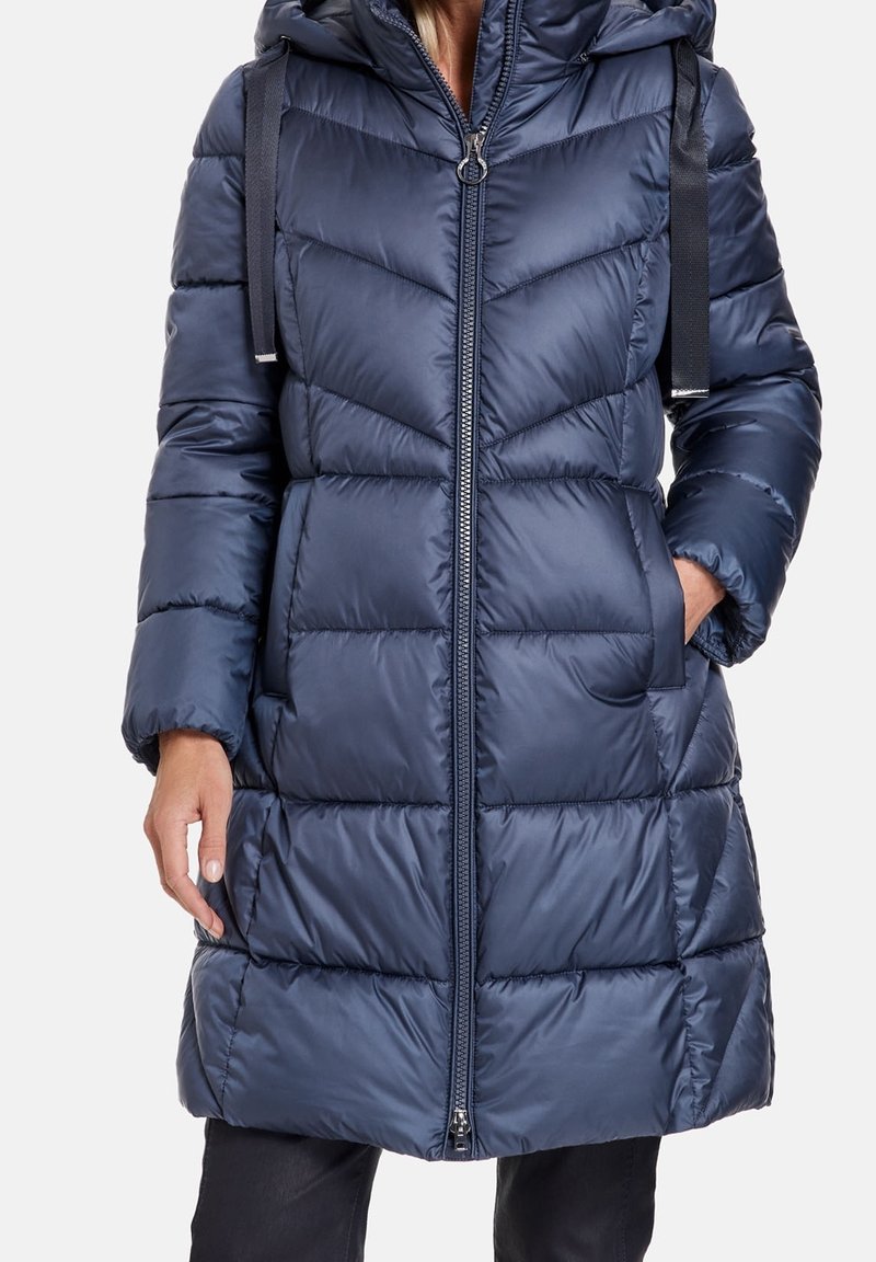 Polstret frakke i marineblå med hætte, chevronmønster, lynlås foran og sidelommer. Fremstillet af skinnende, glat stof med justerbare stropper.