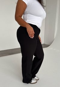 Black tailored pants with a smooth texture, featuring side pockets, paired with a fitted white t-shirt and silver-accented sneakers.