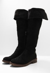 Czarne zamszowe kozaki za kolano z podwiniętym mankietem, bocznym zamkiem i teksturowaną brązową podeszwą. Charakteryzują się minimalistycznymi metalowymi elementami i gładkim wykończeniem.