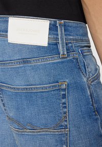 Jeans din denim albastru cu o etichetă din piele albă inscripționată "JACK & JONES". Cusături galbene vizibile și un design clasic cu cinci buzunare.